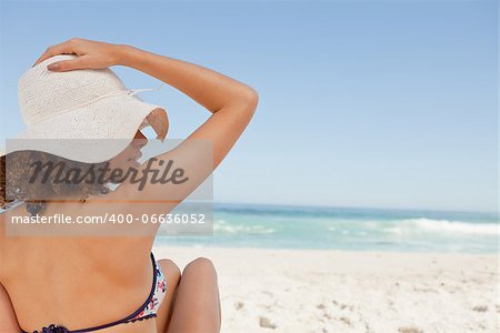 Young woman sitting in front of the ocean while holding her hat on her head