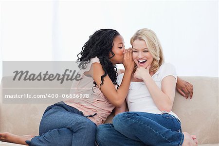 A woman sitting on a couch is whispering into her smiling friends ear