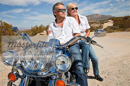 Senior couple wear sunglasses seated on motorcycle on desert road