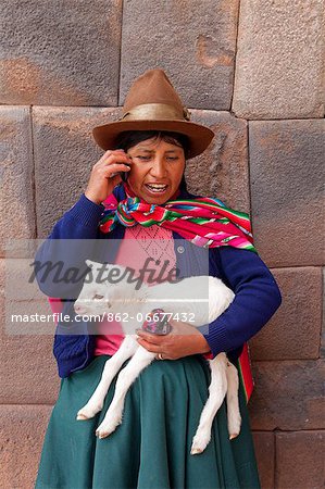 South America, Peru, Cusco. A Quechua woman standing in front of an Inca wall, holding a lamb and wearing a bowler hat and a liclla    while talking on a cell phone in the UNESCO World Heritage listed former Inca capital of Cusc