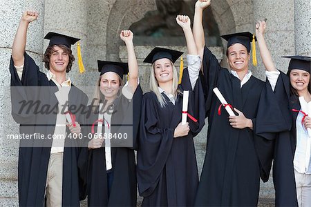 Smiling graduates posing while raising arms in front of the university