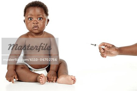 An infant sitting in front of a white background, while a hand holds a syringe.