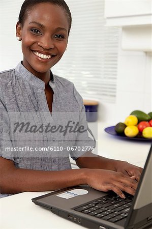 Young African woman working on a laptop in the kitchen, smiling at camera