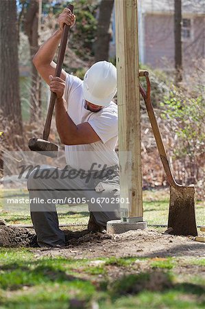 Hispanic carpenter using sledge hammer to tamp dirt at deck footing