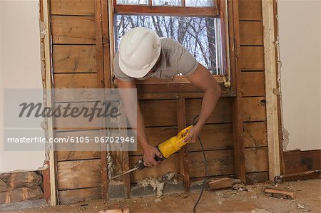 Hispanic carpenter using reciprocating saw to cut wall frame through window access