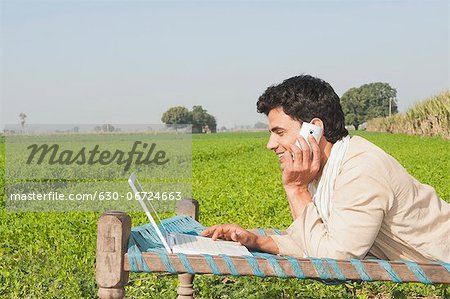 Farmer using a laptop and talking on a mobile phone in the field, Sonipat, Haryana, India