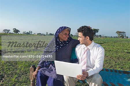 Businessman sitting in the field near his mother and using a laptop, Sonipat, Haryana, India