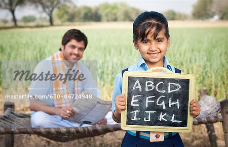 Schoolgirl showing a slate with his father sitting in the background, Sohna, Haryana, India