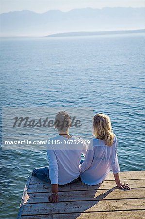 Croatia, Young couple sits on boardwalk, rear view, by the sea