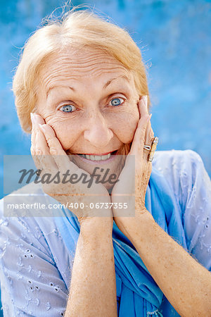 Old blonde woman portrait, lady in elegant clothes with hands on face for joy and surprise on blue background