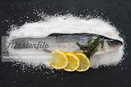 Photo of a full raw fish on a bed of sea salt and on black slate with lemon slices and thyme.