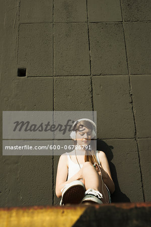 Young woman sitting on ground, leaning against cement wall, listening to MP3 player