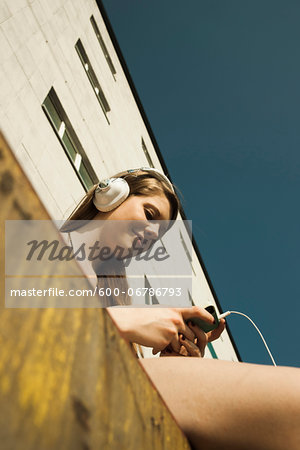 Young woman sitting outdoors, next to building, listening to MP3 player