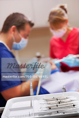 Dental cleaning tools on a white tray in the foreground. Male dentist and female dental hygienist working on patient, blurred in the background.