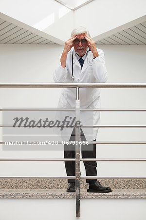 Stressed doctor is leaning on the railing with hands on his forehead while in hospital corridor