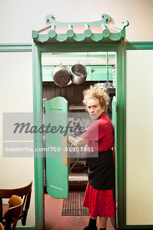 Waitress with curly red hair holding several plates and looks over her shoulder as she walks through swinging double doors into the kitchen of a restaurant in California.