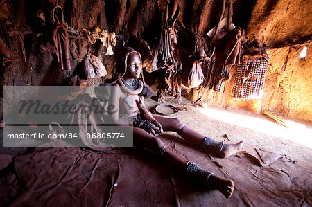 Young Himba woman inside a traditional mud dwelling hut wearing traditional dress and jewellery and with her skin covered in Otjize, a mixture of butterfat and ochre, Kunene Region, formerly Kaokoland, Namibia, Africa