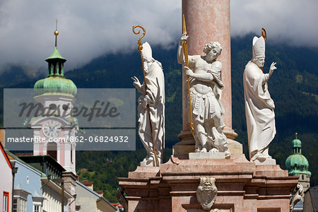 Austria, Tyrol, Innsbruck. Detail of religious figures on a column in the historical centre with a church tower and the alps in the background surrounded by mist