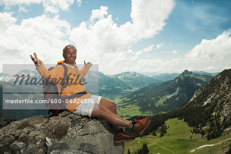 Mature man sitting on cliff with arms raised in air, hiking in mountains, Tannheim Valley, Austria Mature man sitting on cliff with arms raised in air, hiking in mountains, Tannheim Valley, Austria