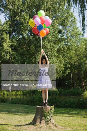 Young woman holding bunch of balloons, standing on tree stump