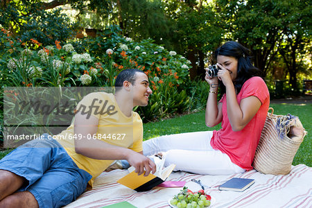 Young couple sitting on picnic blanket, woman photographing man