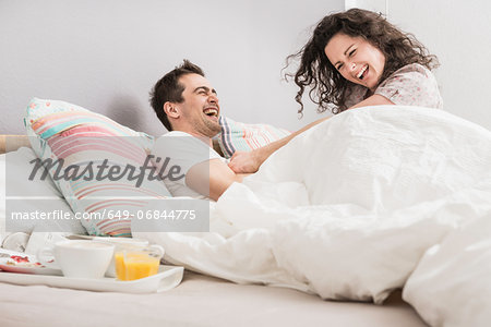 Mid adult couple lying in bed, breakfast on tray, pillow fight
