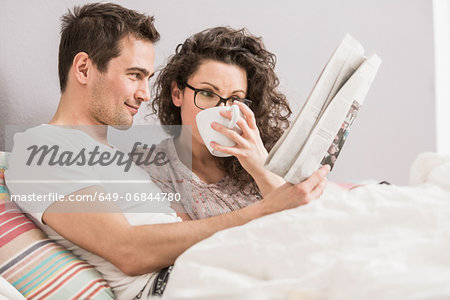 Mid adult couple reading newspaper in bed, drinking coffee