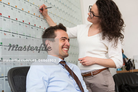 Woman writing on wall calendar, man sitting on office chair