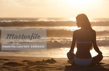 A relaxed sexy young brunette woman or girl wearing a bikini sitting on a deserted tropical beach at sunset or sunrise