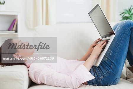 Side view of casual young woman lying on sofa and using laptop