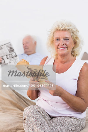 Smiling aged couple reading book and newspaper in the bedroom