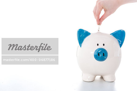 Hand putting coin into blue and white piggy bank with copy space on white background