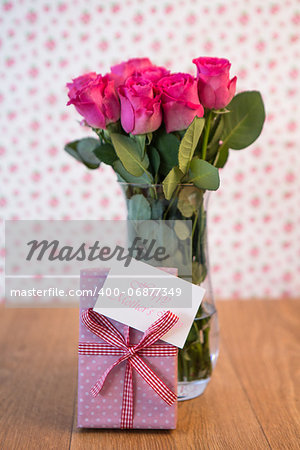 Bunch of pink roses in vase with pink gift leaning against it and mothers day card on wooden table