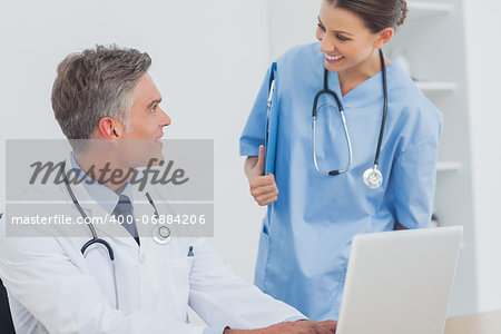 Doctor with a clipboard talking to a colleague while he is using a laptop