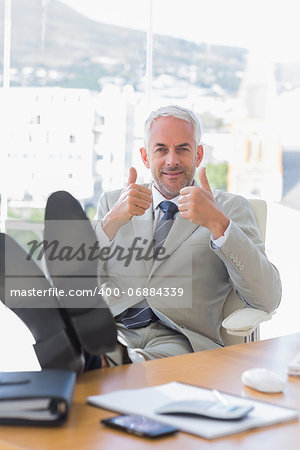 Happy businessman giving thumbs up with feet up on his desk