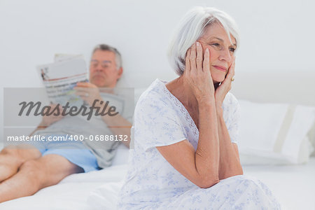 Anxious mature woman sitting on bed while husband is reading a newspaper on the background