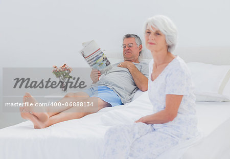 Mature woman sitting on bed while husband is reading a newspaper on the background