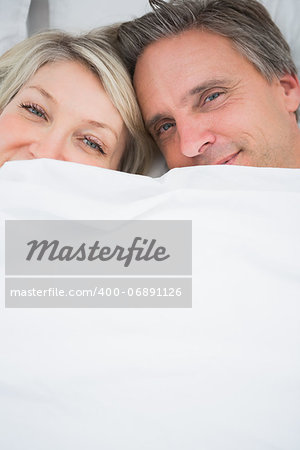 Couple smiling at camera from under the covers at home in bed
