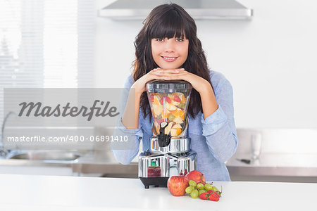 Pretty brunette leaning on her juicer full of fruit smiling at camera at home in kitchen