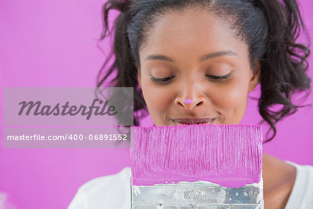 Cheerful woman holding paintbrush with paint on her nose against a pink wall