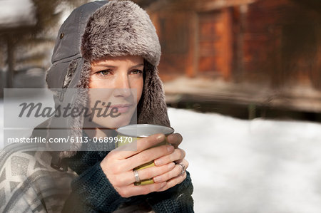 Pensive woman in fur hat drinking coffee outside cabin