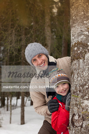 Portrait of happy father and son behind tree trunk in snowy woods