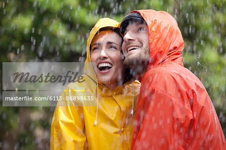 Happy couple in raincoats looking up at rain