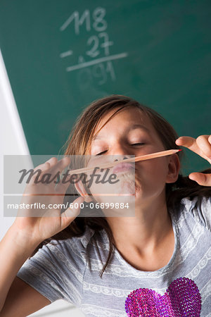 Portrait of girl standing in front of blackboard in classroom, holding pencil over mouth, Germany