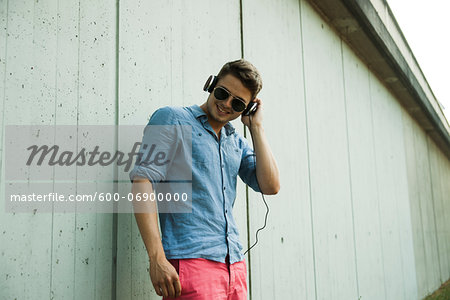 Young man standing next to wall of building outdoors, wearing headphones and sunglasses, Germany