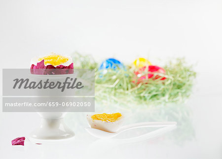 A boiled egg with the top cut off, in an eggcup, with an Easter nest of colourful eggs in the background