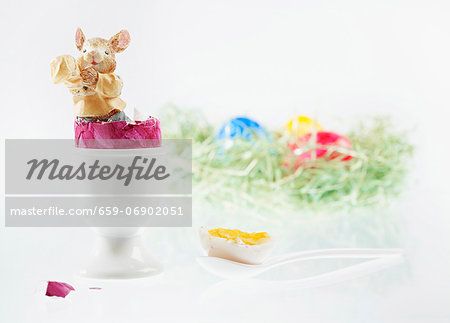 An Easter bunny figurine in a boiled egg with the top cut off, with an Easter nest of colourful eggs in the background