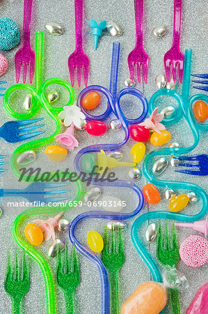 graphic overview of coloured plastic party forks and blue and purple curved drinking straws on a silver glittery background with sweets