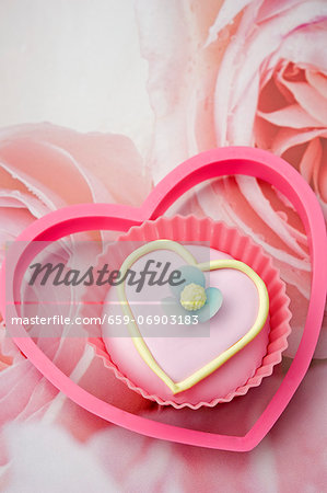 close up of one pink iced, love heart shaped cup cake with a pink plastic heart shape biscuit cutter