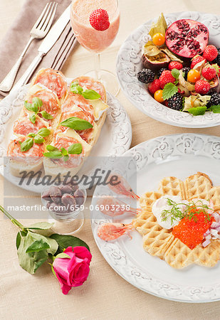 A breakfast of a caviar-topped waffle, slices of bread topped with cheese and ham, fresh fruits and a strawberry and mango smoothie